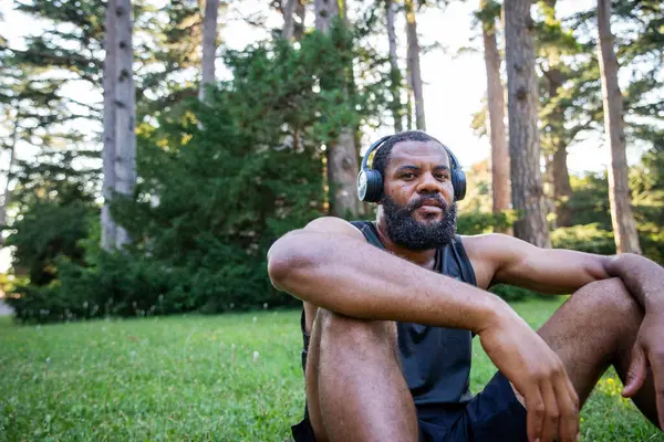 An African Athlete wearing headphones sits in a grassy field. He is wearing a black tank top and shorts
