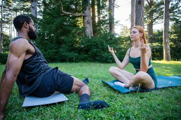 A man and a woman are sitting on the grass in a park. The man is wearing a black tank top and shorts, while the woman is wearing a green top and shorts. They are both practicing yoga poses