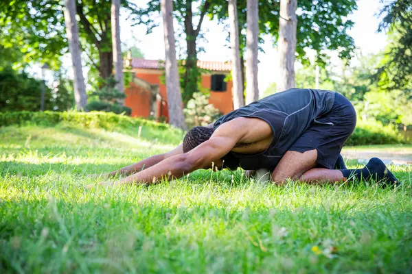 A man is doing a yoga pose in a grassy field. The man is bent over and he is in a relaxed state. The field is lush and green, and there are trees in the background