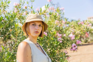 Hasır şapkalı ve beyaz elbiseli bir kadın çalıların önünde kendinden emin bir bakışla dikiliyor..