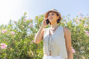 Pembe çiçek tarlasında dikilen bir kadın cep telefonuyla konuşuyor. Hasır bir şapka ve beyaz bir gömlek giyiyor.