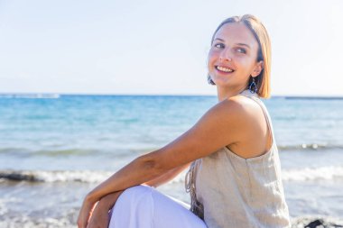 Bir kadın okyanusun kenarındaki bir kayanın üzerinde oturuyor, gülümsüyor ve denize bakıyor.