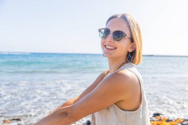 Bir kadın okyanusun yakınındaki bir kayanın üzerinde otururken gülümsüyor ve güneş gözlüğü takıyor..