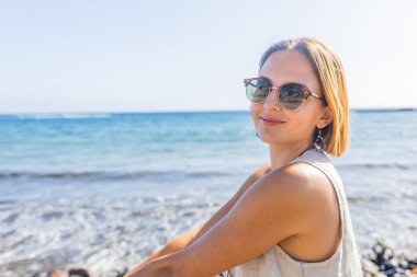 Yaz boyunca plajda oturan bir kadın güneş gözlüğü takmaktan mutlu olur..