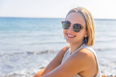 Bir kadın kumsalda otururken gülümsüyor ve güneş gözlüğü takıyor. Rahatlama kavramı