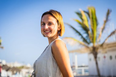 Bir kadın gülümsüyor ve bir palmiye ağacının önünde duruyor, güneşli bir günde pozitif bir atmosfer yaratıyor..