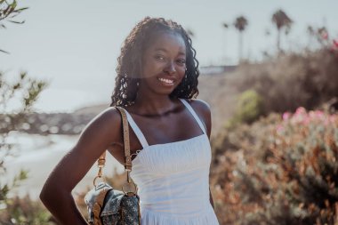 Uzun saçlı siyah bir kadın gülümsüyor ve elinde bir çanta tutuyor.