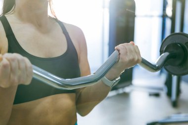 Bir kadın spor salonunda halter kaldırıyor. Siyah bir atlet ve beyaz bir bileklik giyiyor.