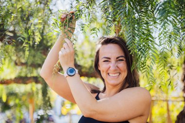 Bir kadın gülümsüyor ve elinde bir dal tutuyor. O bir saat ve siyah bir atlet giyiyor.