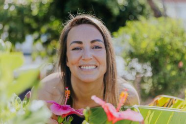 Bir kadın gülümsüyor ve çiçek bahçesinin önünde poz veriyor. Çiçekler pembe. Manzara neşeli ve rahat..