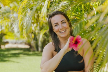 Bir kadın elinde pembe bir çiçekle poz veriyor. Gülümsüyor ve anın tadını çıkarıyor. Mutluluk ve rahatlama kavramı, kadının doğa ve güzel çiçeklerle çevrili olması gibi.