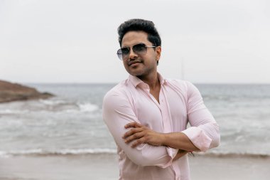Güneş gözlüklü, kolları katlanmış, kendine güvenen adam okyanusun yanında duruyor..