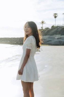 Plajda beyaz bir elbise giymiş bir kadın var. Gülümsüyor ve kameraya bakıyor. Plaj güzel ve sakin bir yerdir. Arka planda okyanus vardır.