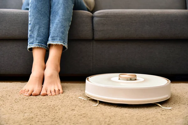 Modern robot vacuum cleaner near sofa with woman legs in room. Housewife feet. Robot vacuum cleaner cleaning carpet.