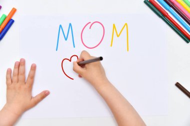 Kids girl draws the word mom on a paper sheet with markers and drawing a postcard for mom, top view. Sincere love of parents. Preparing for the holiday in  clipart