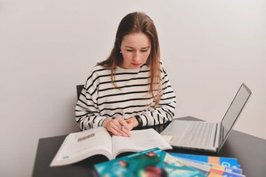 Konsantre bir kadın kitaba bakıyor, çalışıyor ya da çalışıyor, bir masada dizüstü bilgisayarla oturuyor, defterler, kitaplar, yine gri duvar arka planı, yüze odaklanıyor..