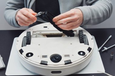 Odaklanmış adam bir robot elektrik süpürgesi üzerinde çalışıyor, bileşenlerini dikkatlice tutuyor. Onarım işlemi masadaki birden fazla aracı içerir.