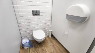 Duvara monte edilmiş tuvaleti olan modern umumi tuvalet, beyaz geometrik fayanslar, bir tuvalet kağıdı makinesi ve bir çöp kovası. Ahşap döşemeli temiz ve minimalist tasarım.