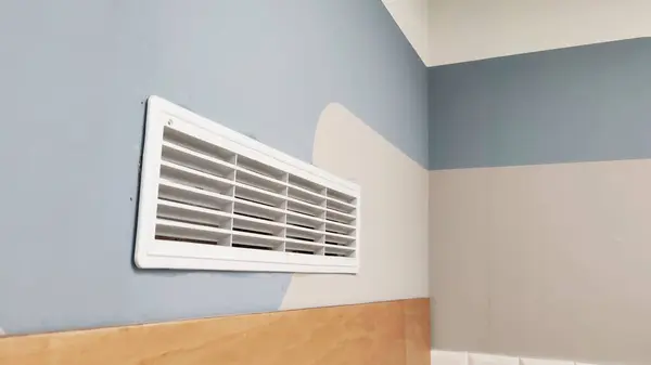 Beyaz duvar havalandırma ızgarası mavi, bej ve gri geometrik desenli renkli bir duvara monte edilmiş. Havalandırma boşluğu odada uygun hava akımını ve havalandırmayı sağlar..