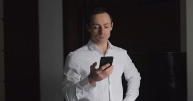 Klasik tişörtlü bir iş adamı pencerenin yanında telefonla konuşur ve saate bakar.