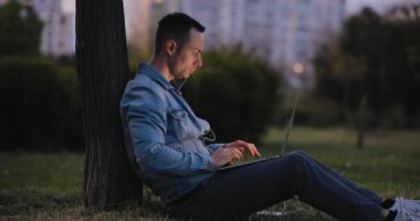 Dizüstü bilgisayarla çalışan bir adam parkta bir ağaç gövdesine yaslanıyor.