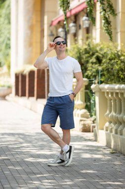 Şortlu ve tişörtlü yakışıklı adam şehir sokaklarında güneş gözlüklü poz veriyor.