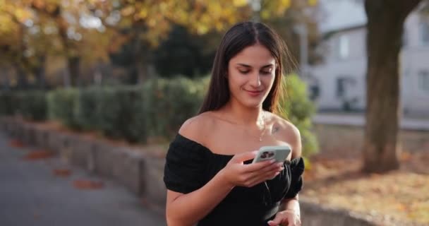 秋の路地で携帯電話を使用してゆっくり動くかなり笑顔の女性 — ストック動画