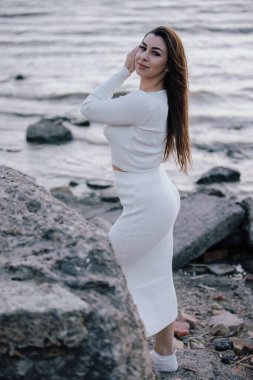 Deniz kenarındaki taşlara poz veren beyaz elbiseli zarif bir bayan.