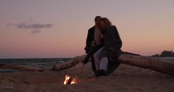 カップルは夕方にビーチで火の近くに優しさと一緒に座っています — ストック動画