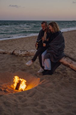 Akşamları kumsalda, ateşin yanında şefkatle oturan çift.