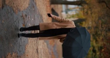 Palto giyen bir adam, sonbahar geçidi boyunca şemsiye altında ağır çekim dikey video ile yürüyor.