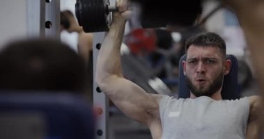 Spor salonunda aynanın yanında dambıllarla kasları çalıştıran bir adam.