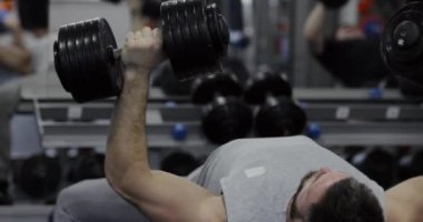 Güçlü bir adam spor salonundaki bankta duran dambıllara basar.