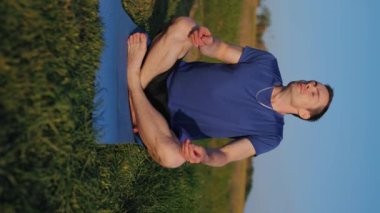 Nilüfer yogası yapan bir adam doğada bir mindere oturmuş, dikey video çekiyor.