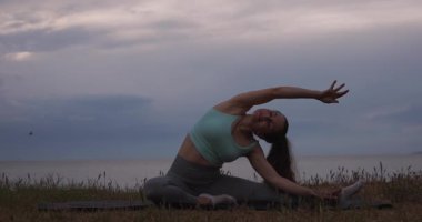 Yoga yapan bir kadın tepenin üstünde deniz ve günbatımına karşı egzersiz yapıyor.