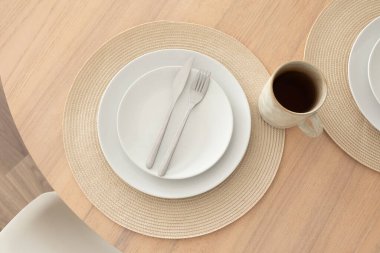 Tahta masada minimalist masa. Beyaz seramik tabaklar, çatal ve bıçakla örülmüş yuvarlak bir altlık ve siyah kahve dolu seramik bardaklar.