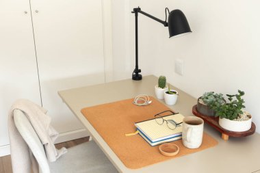 Konforlu ofis sahnesi bej masası, mantar paspası, siyah masa lambası, seramik kahve fincanı, defterler, gözlükler ve küçük saksı bitkileri, sulu bitkiler ve kaktüsler. Concent üretkenliği, uzaktan çalışma, serbest çalışma, farkındalık veya iç tasarım