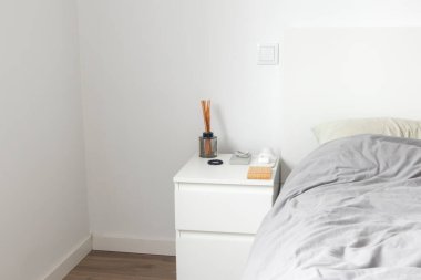 Minimalist estetik yatak odasının içi. Beyaz komodinli komodin, şarj kablosu, kozmetik kablosu, telefon standı, kablosuz şarj aleti ve saç teli.