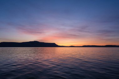 Turuncu, sarı, pembe, mor ve kırmızıdan oluşan güzel bir şafak gökyüzü hafif dalgalı okyanus suyunda yansıyor Batı İzlanda dağları ise bir siluet gibi duruyor..