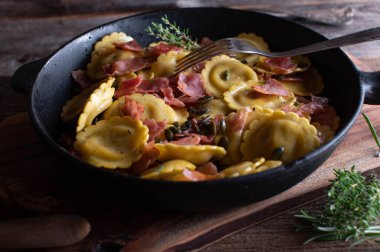 Mantarlı Ravioli, İtalyan jambonu, tereyağı ve kabak çekirdeği. Ahşap bir masada demir bir tavada servis edilmiş.