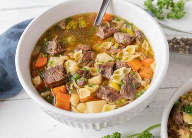 Delicious and healthy beef noodle soup with clear broth, root vegetables and noodles. Served in a white large bowl with ladle on kitchen table. Closeup and top view