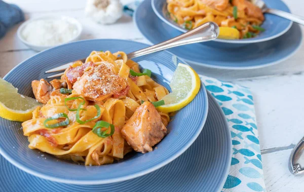 Delicious salmon dish with tagliatelle and tomato cream sauce. Topped with grated parmesan cheese and served with garnish on blue dishes on kitchen table background. Front view and closeup.