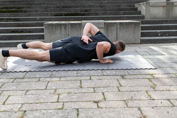 Advanced push-up variation with one arm push-up is doing by a young athletic man outdoors on concrete background. Front view