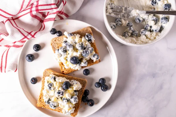Delicious and healthy high protein breakfast toast with low fat cottage cheese, blueberries and honey on a plate on light kitchen background.