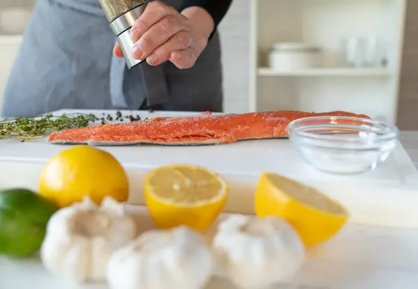 stock image Woman with apron seasoning a raw half salmon fillet with a pepper shaker on a cutting board in the kitchen