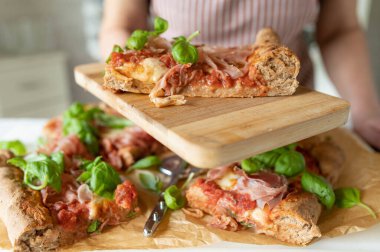 Hand holding or serving a fresh piece of healthy whole wheat pizza with ham, mozzarella cheese, tomatoes and basil leaves