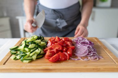 Elinde bıçak, ellerinde salatalık, domates ve soğan gibi sebzeler olan bir kadın.