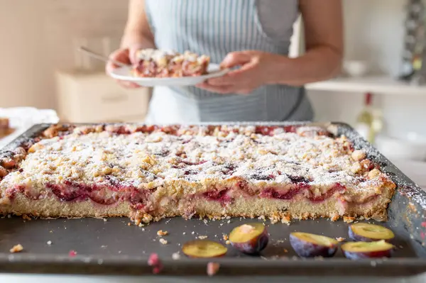 stock image German plum cake with streusel served by a woman with apron in the kitchen. Homemade sheet cake. Closeup, front view in the kitchen