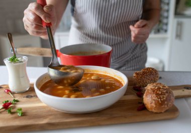 A woman with apron pouring healthy bean soup on a plate in the kitchen clipart