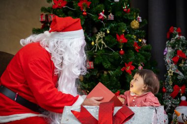 Beyaz bebek Noel Baba 'dan hediye alıyor. Noel kutlaması konsepti için süslenmiş Noel ağacının yanında.